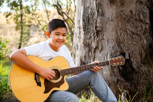 Boy playing country music on acoustic guitar in bushland - Australian Stock Image