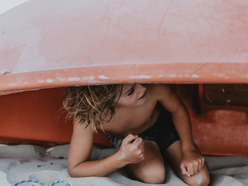Boy playing hide and seek under red boat on beach - Australian Stock Image