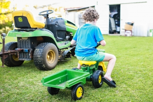 Boy playing on ride on tractor with trailer next to ride on lawn mower - Australian Stock Image