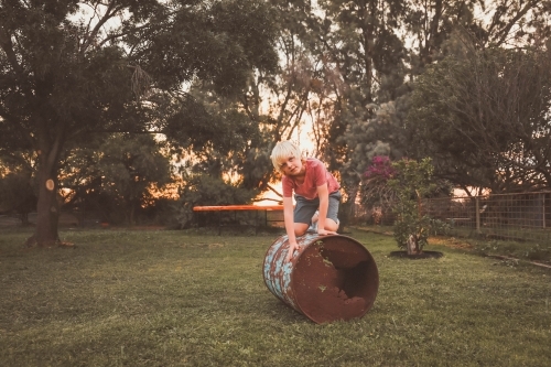 Boy playing on rusty metal drum in country backyard on farm - Australian Stock Image