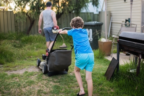Boy pushing lawn mower mowing lawn with father holding whipper snipper - Australian Stock Image
