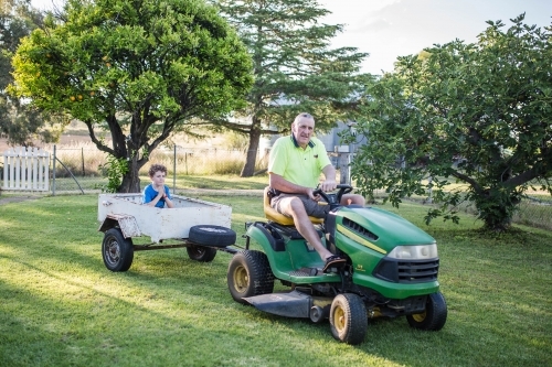 Boy sitting in white trailer being towed by grandfather on ride on lawn mower - Australian Stock Image