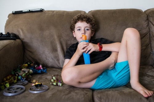 Boy sitting on lounge with toys eating ice block - Australian Stock Image