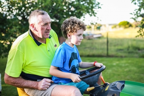 Boy sitting on ride on lawn mower with grandfather - Australian Stock Image