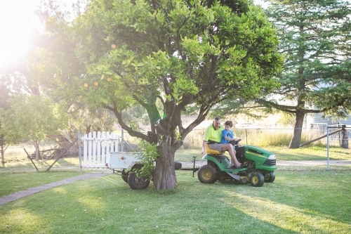 Boy sitting on ride on lawn mower with grandfather towing white trailer - Australian Stock Image