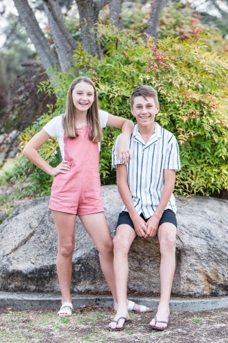 Boy sitting on rock in garden while sister leans on his shoulder smiling - Australian Stock Image