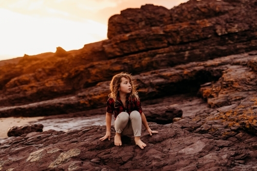Boy sitting on rocks at the ocean at sunset - Australian Stock Image