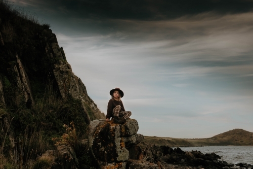 Boy sitting on rocks next to cliffs and ocean a very dark moody sky - Australian Stock Image