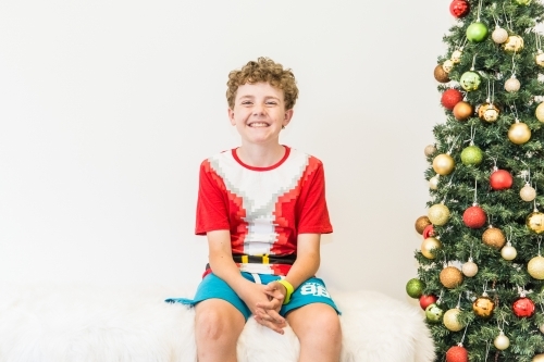 Boy sitting on rug next to Christmas tree smiling excited - Australian Stock Image