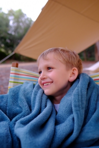 Boy smiling, sitting outside tent in camping chair - Australian Stock Image