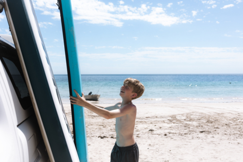 Boy stacking paddle boards against car on clear blue beach in full summer sun - Australian Stock Image