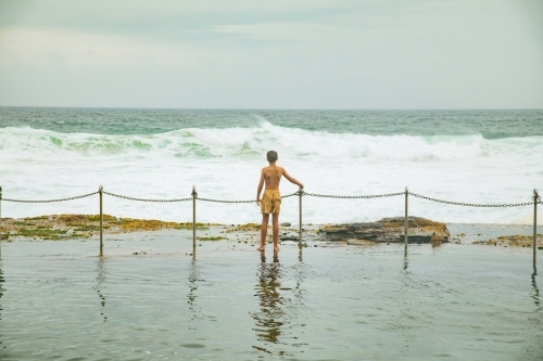 Boy standing at the fence on the edge of the Bogey Hole in Newcastle, NSW on an overcast day