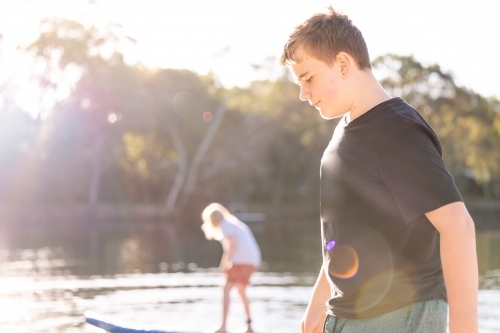 Boy standing in golden afternoon sun with brother on SUP in background - Australian Stock Image