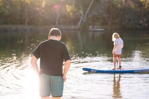 Boy standing in golden afternoon sun with brother on SUP in background