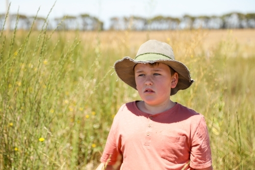 Boy standing in long grass on farm wearing green hat on hot summer day - Australian Stock Image