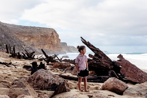 Boy standing on beach next to shipwreck with ocean and cliffs behind
