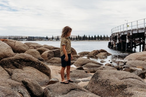 Boy standing on rocks looking at the ocean next to a jetty - Australian Stock Image