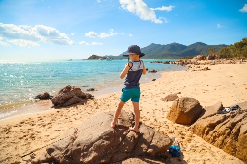 Boy standing on rocks on tropical beach with islands in the distance - Australian Stock Image