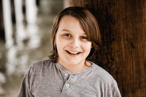 Boy standing under a pier at the beach smiling - Australian Stock Image