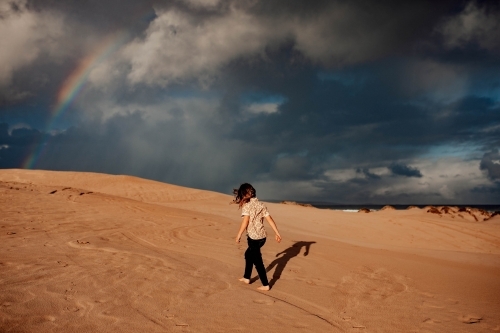 Boy walking on sand dune with rainbow in sky - Australian Stock Image