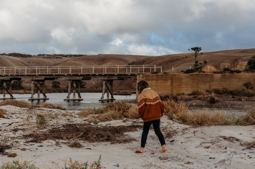 Boy walking on the beach with a bridge and hills in the distance - Australian Stock Image