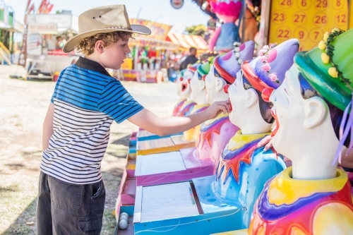 Boy wearing akubra hat placing ball in mouth of clowns sideshow alley at local show - Australian Stock Image