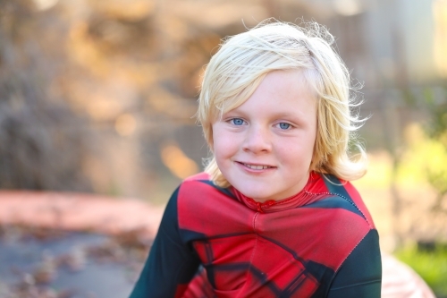 Boy wearing red superhero suit sitting on trampoline covered in autumn leaves - Australian Stock Image