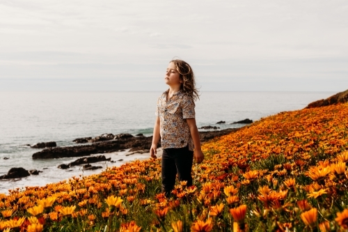 Boy with eyes closed and face to the sun standing in a field of flowers on a clifftop next to ocean - Australian Stock Image