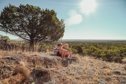 Boys sitting on rock on hilltop with flat landscape and view of the horizon in background - Australian Stock Image