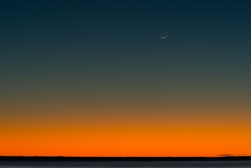 Bright orange and dark blue post sunset sky with crescent moon and low horizon - Australian Stock Image