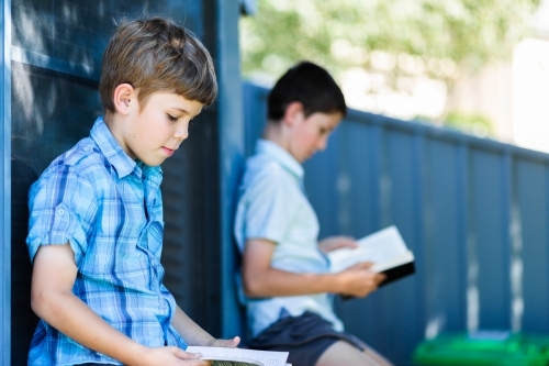 Brothers sitting against window in backyard reading books - Australian Stock Image