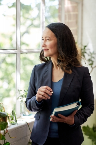Business woman stands at the window of a warehouse style office thinking - Australian Stock Image