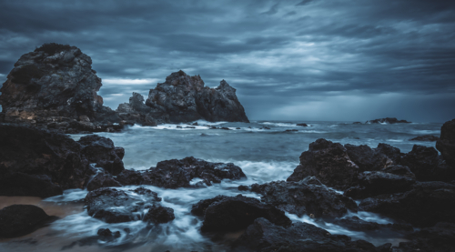camel rock bermagui with dark storm clouds over rocks on ocean shore - Australian Stock Image