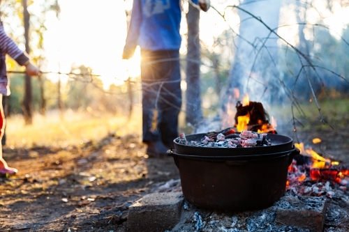 Camp oven over campfire cooking dinner - kids play nearby fire - Australian Stock Image