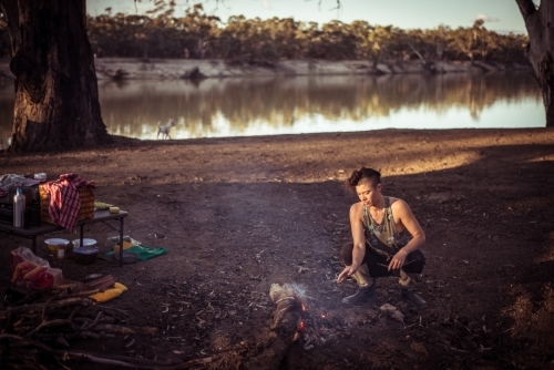 Camping life - person squatted by a campfire next to a river - Australian Stock Image
