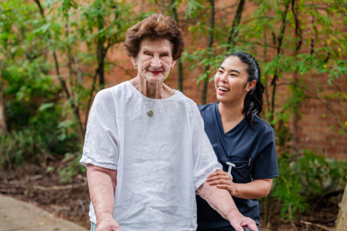 Caregiver in dark blue scrubs assisting an elderly woman on her rollator walking outdoors - Australian Stock Image