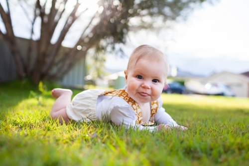 Cheeky baby girl with tongue poking out lying on green spring grass on front lawn - Australian Stock Image