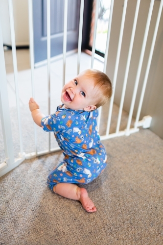 cheeky baby pulling up on baby gate near front door of home - Australian Stock Image