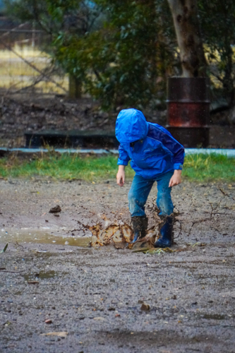 child in blue rain jacket jumping in muddy puddles - Australian Stock Image