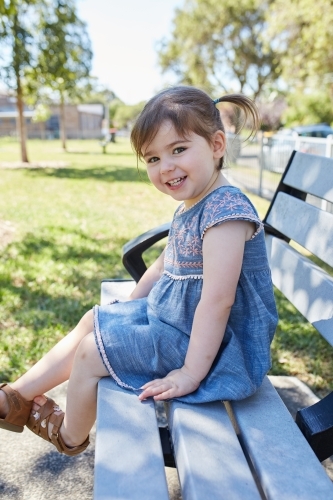Child smiling on park bench wearing a blue dress - Australian Stock Image