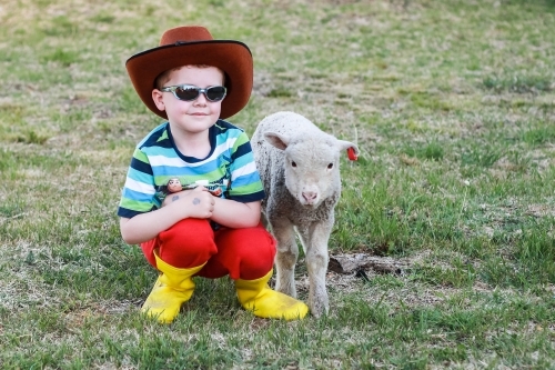 Child wearing akubra hat and sunglasses sitting with lamb - Australian Stock Image