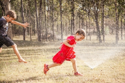 Children running under sprinkler in backyard, Aussie summer childhood - Australian Stock Image