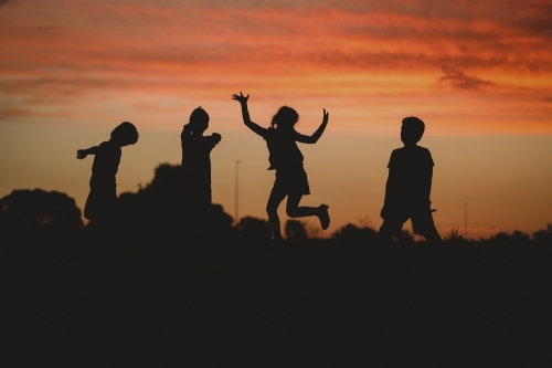 Children silhouette playing at sunset with girl jumping - Australian Stock Image