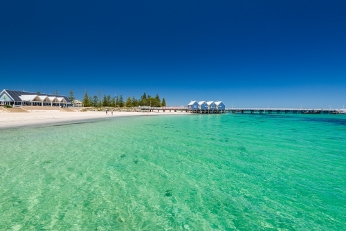Clear green water and a white sandy beach with Jetty in background and clear dark blue sky