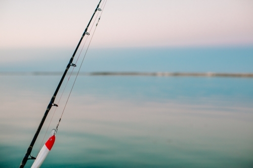 Close-up of a fishing rod with lure with a calm ocean and sky in the background - Australian Stock Image