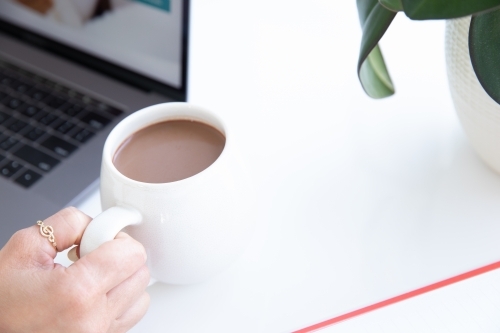 Close up of a ladies hands holding a hot chocolate beside laptop - Australian Stock Image