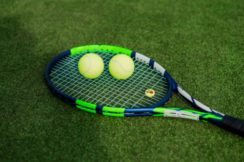 Close-up of a tennis racquet resting on green grass with two yellow tennis balls nearby - Australian Stock Image