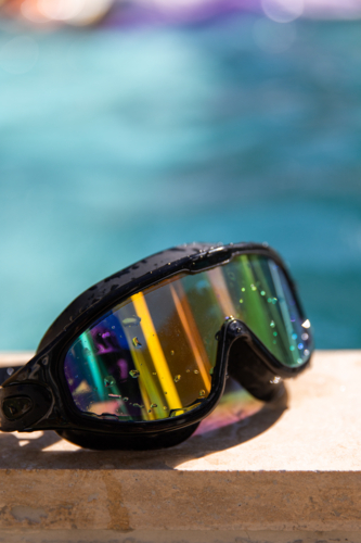 Close-up of black swimming goggles with rainbow-reflective lenses resting on the edge of a pool - Australian Stock Image
