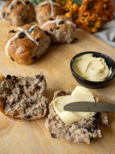 Close up of cut Hot Cross Bun with butter and knife on cutting board with full buns and flowers - Australian Stock Image