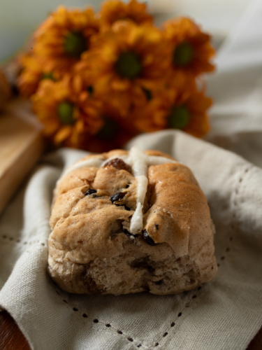 Close up of single Hot Cross Bun on decorative linen napkin with orange flowers in background - Australian Stock Image